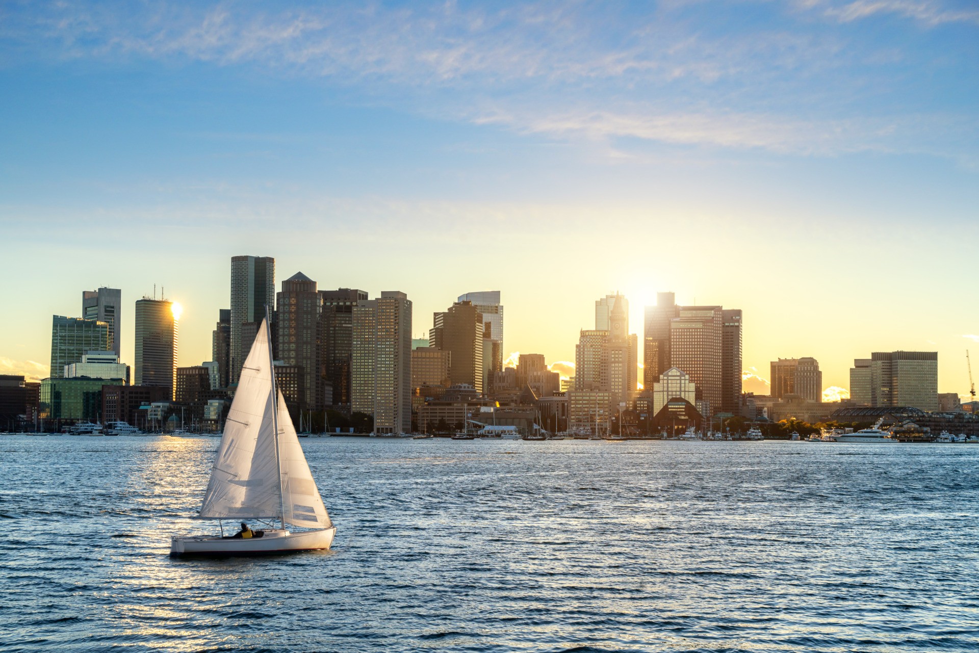 Our Approach. Sailboat sailing at sundown in the Boston Harbor with the downtown skyline in the background.