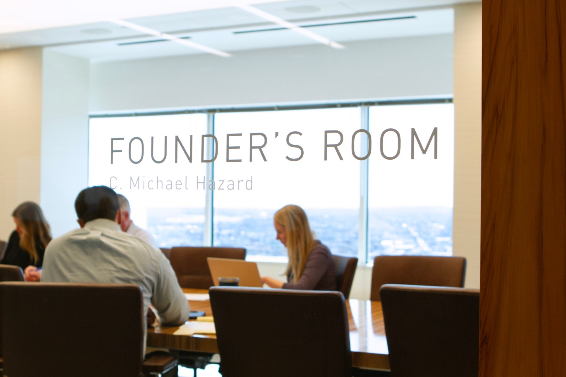 Rooted in growth. Powered by people. Image of team meeting in Westfield's office board room with picture in the back displaying views of the Boston Harbor. The image is shot through the room's glass walls with that have the name etched on the outside spelling out "Founder's Room".